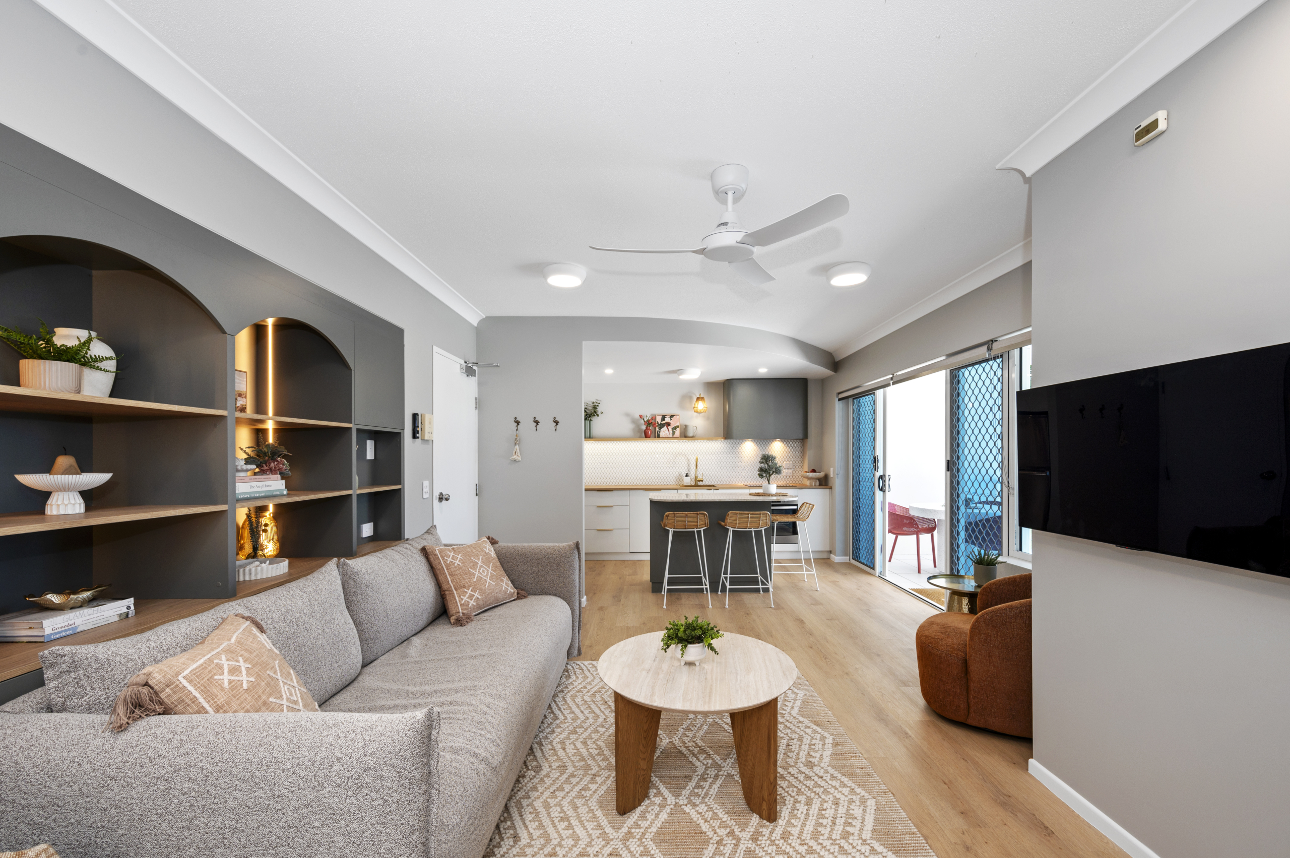 Living area in Koola Palms 101 with grey sofa, open shelving and wall mounted TV looking through to the kitchen